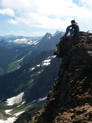 Junger Mann sitzt oben auf einem Berg und hat Aussicht auf weitere Berge