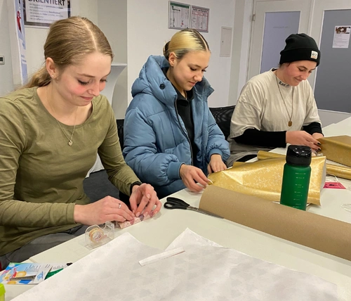 3 Jugendliche sitzen an einem Tisch und packen Geschenke ein.