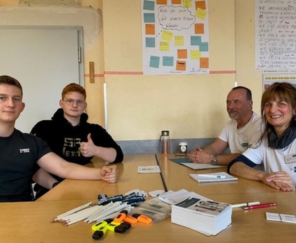 Zwei Erwachsene in einheitlichen T-Shirts sitzen mit zwei Jugendlichen an einem Tisch und haben vor sich Papier, Flyer und Stifte.