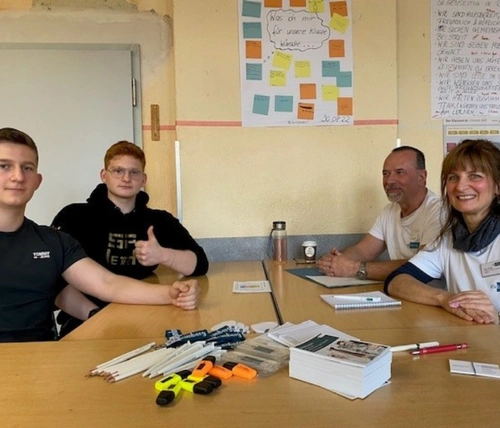 Zwei Erwachsene in einheitlichen T-Shirts sitzen mit zwei Jugendlichen an einem Tisch und haben vor sich Papier, Flyer und Stifte.