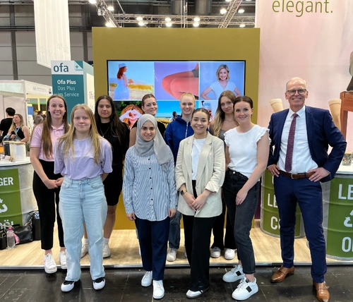 8 Jugendliche stehen zusammen mit ihrer Ausbildungsleiterin und dem Geschäftsführer vor einem Messestand