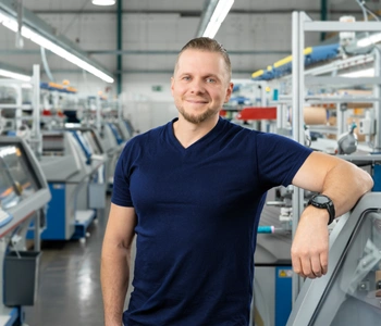 Mann Mitte 30 in einer Halle mit Strickmaschinen, er lehnt an einer Maschine