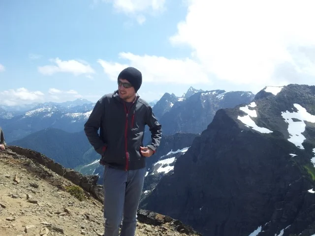 Aufnahme von jungem Mann, der in Jacke und Sonnenbrille vor Bergen zu sehen ist.